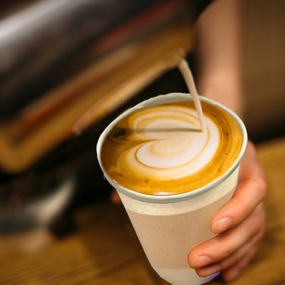 A person pours a latte into a cup. The cup is a 12oz disposable coffee cup with lids and sleeves.