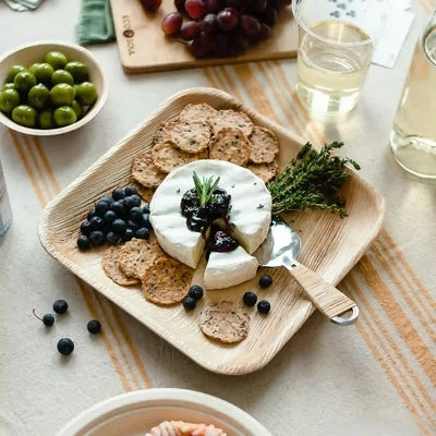 Table with a variety of cheese, grapes, and olives beautifully arranged on 10 Inch Square Compostable Palm Leaf Plates.