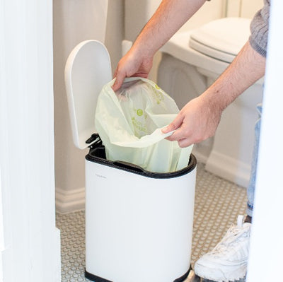 A person placing a trash can in a bathroom with Compostable Small Kitchen Trash Bags on the dustbin.