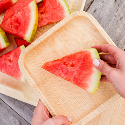 Person holding a piece of watermelon on a 8 Inch Square Compostable Palm Leaf Plates