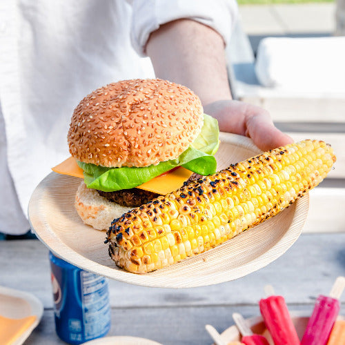 A person holding a plate with a hamburger and corn on it. Plate is made of 10 Inch Round Compostable Palm Leaf Plates.